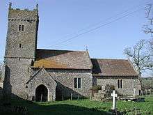 A stone church seen from the south, with a square battlemented tower at the left, then the nave, and a lower chancel at the right. In front of the church is the base of a medieval cross