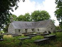 A T-shaped low stone church with a slate roof seen beyond grave slabs