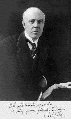 A balding, white-haired man wearing 1920s formal business attire (jacket and waistcoat with wing collar and tie) leans to his left on a table and looks directly at the viewer. His right hand rests in front of his left on the edge of the table next to some papers and a pen.