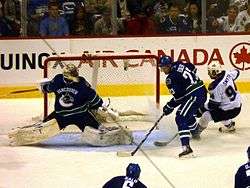 An ice hockey goaltender stretches across the net to get into position as a teammate stands to the right of the net, keeping an opposing forward away.