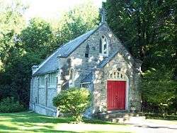 A light brown stone chapel with a projecting front, red door and steep roof with a cross on top