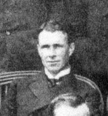  A man, fresh-faced with dark, brushed-back hair, seated among a group. He is wearing a naval officer's uniform with a high, stiff collar