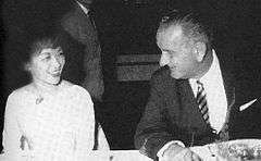 A middle-aged lady wearing a light-coloured dress and with short hair, fluffy at the front, sits at a dinner table smiling. To the right is a taller, older man in a dark suit, striped tie and light shirt who is turning his head to the left, talking to her. A man in a suit is visible, standing in the background.