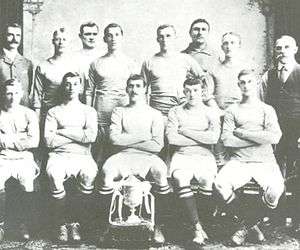 A group of thirteen men, eleven in association football attire typical of the early twentieth century and two in suits. A trophy sits in front of them