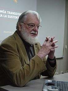 Manfred Max Neef sits at a table near a notebook computer. On the wall behind him is a slide from a presentation.