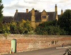 Yellow stone house with tall chimneys behind brick wall.