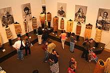 Museum goers look an exhibit of clothing from New Zealand's Toi Maori people. On the wall behind are photos of individuals wearing examples of the clothing.