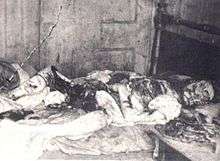 Black and white photograph of an eviscerated human body lying on a bed. The face is mutilated.