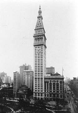 Aerial view of a thin, 50-story building with a light exterior; the building has a pyramidal roof with a large spire, and a clock is visible below the roofline.