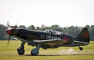 Side view of a piston-engined fighter painted in green and brown with large red star insignia taking off. A slogan in Cyrillic letters is painted on its side.