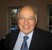 An elderly balding man with some thin white hair, wearing clear-rimmed glasses, a dark suit and blue tie, smiling.