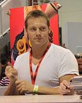 A mid-aged man with short brown hair, is sitting behind a table with a pen in his hand, and is looking to his left.