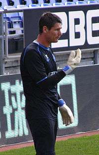 Dark-haired white man wearing tracksuit and bulky gloves stands at the edge of a sports field.