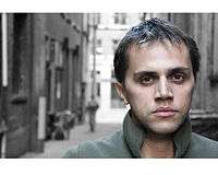 A mixed male with light brown hair, hazel eyes and a clean shaven face, in front of an alley. He is wearing a green jacket.