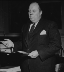 Perrow standing at desk motioning with right hand and holding paper in left hand