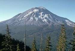 Large volcanic mountain deformed by large cracks and bulges on its slopes, seen from a ridge across a thickly forested valley. A summit crater is partially seen.