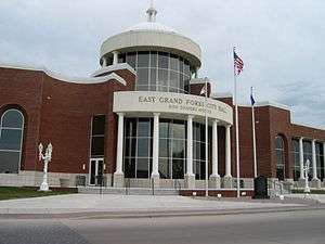 A red brick building with a glass lobby. A memorial sign and flags are located in front of the building.