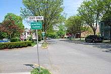 Ground-level view of a road on a sunny day; the street is lined with newly-budding trees.