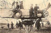 An airship gondola is shown in close-up from mid-left to mid-right; various items of equipment hang from the gondola's side; below the gondola a pyramid of steel tubing protrudes, shielding a cylindrical fuel tank; four crew members are in the gondola; above it mooring ropes and a rip-line are visible. A flat-ended propeller is also visible on the right