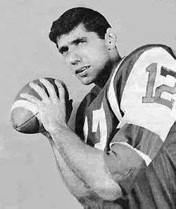 Black and white publicity still of Namath, in pads and jersey with no helmet, holding football over his shoulder as if to throw it (1965).