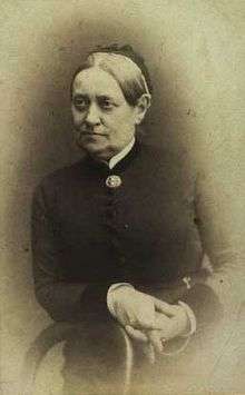 Black and white photo of a woman leaning with her right forearm on the back of a wooden chair