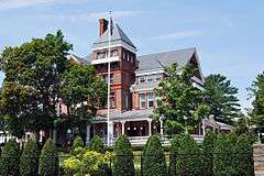 An ornate brick house with a pyramid-roofed tower on the front seen from slightly below. In front is a flagpole and some tall trees, with shrubbery and a chain link fence at the bottom of the image, closer to the camera.