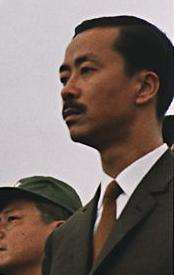 Middle-aged man with side-parted black hair and moustache, in a black suit, white shirt and brown tie. To the left is a clean-shaven Asian man with black hair and a green military cap.