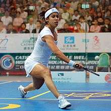 A young female squash player in white, stretching out to get her racket under a low ball, on a blue court with yellow markings, in front of a large, out-of-focus crowd behind glass.