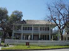 A gambrel-roofed house with large windows, wraparound balcony and first floor porch with Grecian columns. Sashed windows are framed by shutters.