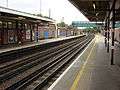 A grey-tiled railway platform bordered by red-bricked walls supporting overhanging roofs from which elongated electrical lights hang