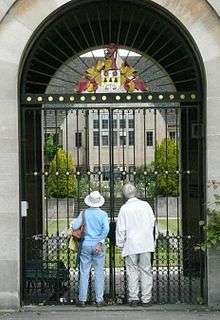 A large gate of metal bars, within a curved arch of light-coloured stone. Two people in casual clothes look through the gate, through which there is a grassed lawn with a pond, steps at the far end and a stone building with large rectangular windows. On the top of the gate, a shield with a coat of arms surmounted by a helmet.