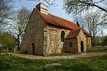 A honey-coloured stone church with a red tiled roof seen from the southwest. On the west end is a window above a door, on the corner is a buttress, a porch in on the south side, and on the roof is a small wooden belfry