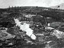 A black and white photograph of the lower sections of a sloped dam, with cranes and other construction equipment on top