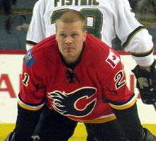 An ice hockey player stands facing the camera bent over at the waist, looking off into the distance. He is wearing a red jersey with black sleeves below the elbows. The jersey's crest is a stylized all black "C" made to look as if the curved part of the "C" has caught on fire. The shoulders have small patches on them, the player's left shoulder is the flag of Canada, on the players right is the flag of Alberta.