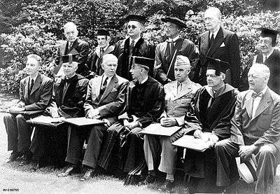 A group of men in uniforms, suits and academic dress sit for a formal group photograph