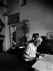 A black-and-white picture of a man seated at a table; an artist's studio is visible behind him