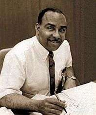 portrait seated at his desk in shirtsleeves and tie with pen in his hand