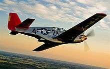 A single nosed-propeller plane is in mid flight above land.  The red nose faces right and the underside of the plane is slightly in view.  The plane has black propellers, grey base paint, and black letters reading A42.  The 4 and 2 are separated by a United States roundel in black with a central white star.  The roundel is also visible on the tops of the planes wings.  The full length of the right side of the plane is visible.