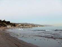 Photograph of a beach. Image of a beach. In it, a pier and several houses can be appreciated.