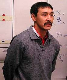 "A colour photo of Paul Okalik standing in front of a chalkboard wearing a grey sweater"