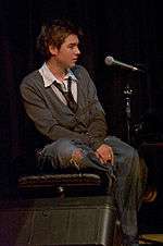 A man sits on a stool next to a microphone