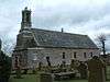 A grey stone chapel, with angular corners. An ornate bell tower is situated on the west of the building.