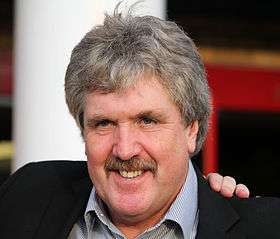 A headshot of a grey haired Caucasian man with a moustache wearing a suit.