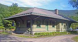 A green building with wooden siding and a peaked black roof viewed at a three-quarters angle. Behind it are railroad tracks and a high wooded ridge.