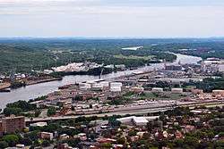 Aerial view of an industrial zone; large silos, cranes, storage tanks, and a highway are seen.