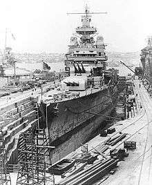 A raised warship in a drained dock surrounded by construction equipment