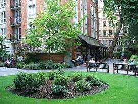 Circle of green grass about 10 yards in diameter, with a roughly 3 yard brown central area containing low bushes. Outward-facing park benches are at the circle's rim, and a multistorey brick building with an awning is in the background, across a sidewalk.