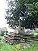 A square platform of three stone steps supporting a carved cross base with a modern octagonal shaft, in the shade of a yew tree