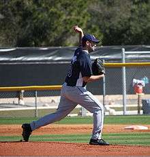 A man throwing a baseball wearing a navy-blue baseball jersey and cap and gray baseball pants