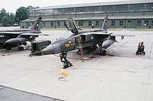 Two dark green jet aircraft (one partly obscured) parked on concrete ramp in front of building.